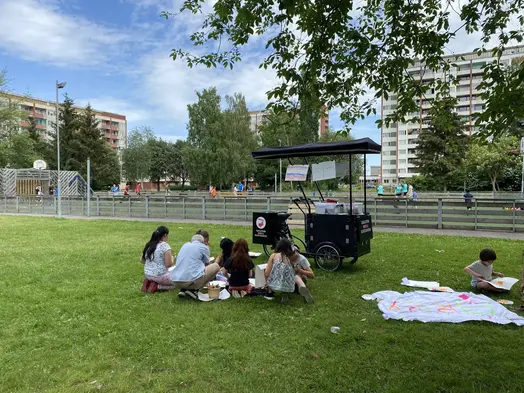 På en gräsmatta står en svart lådcykel, det är Eskilstuna mobila konstmuseum. Framför cykeln, i gräset, sitter människor på huk och på knä och skapar. I bakgrunden syns en basketplan och höga hyreshus.